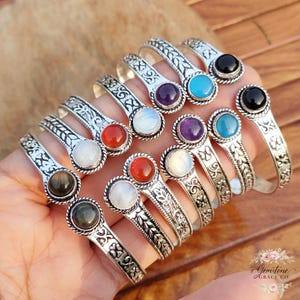 Puede incluir: Una colección de pulseras plateadas con diferentes piedras preciosas. Las pulseras presentan intrincados diseños florales y geométricos. Las piedras preciosas incluyen negro, morado, azul, blanco, rojo y verde. Las pulseras se muestran en una mano.