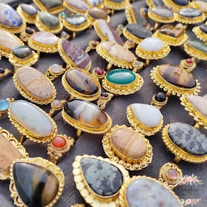 May include: A display of gold rings, each adorned with a different gemstone. The rings showcase a variety of stones in brown, green, white, and black, set in decorative gold mounts. The gemstones are oval, teardrop, and rectangular.