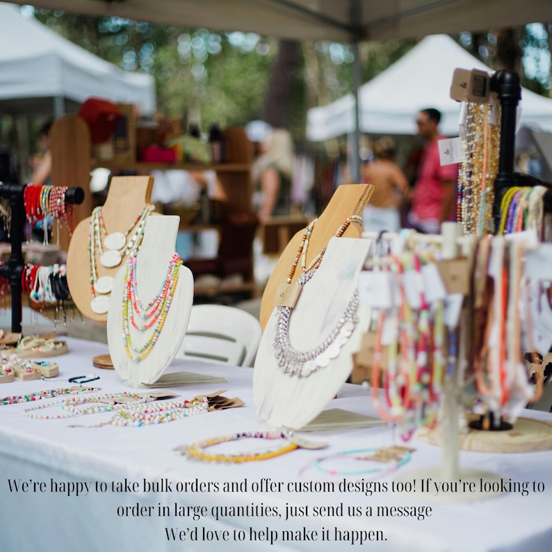 May include: A display of necklaces and bracelets on a white table at an outdoor market. The necklaces are on wooden display stands, showcasing various styles with colorful beads and metallic accents. The text at the bottom says, "We're happy to take bulk orders and offer custom designs too!"