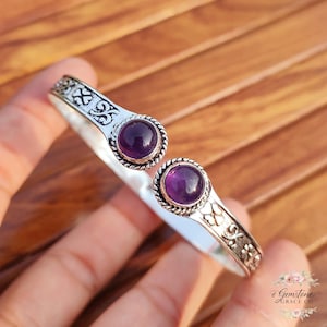 Pode incluir: Uma pulseira de punho prateada com duas pedras preciosas redondas, roxas escuras. A pulseira tem um design aberto com padrões decorativos florais e de rolagem. As pedras são definidas em uma moldura em forma de corda.