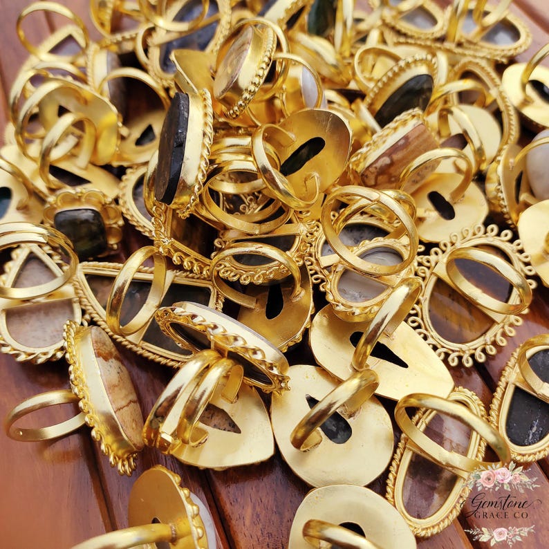 May include: A collection of gold-colored rings with various gemstone settings. The rings feature different shapes, including oval, teardrop, and rectangular designs. The gemstones are in shades of brown, black, and white. The rings are displayed on a wooden surface.