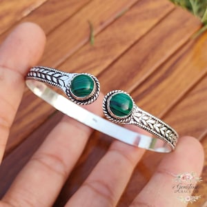 May include: A silver cuff bracelet featuring two round, polished malachite stones. The bracelet has an open design with decorative leaf patterns and rope-like detailing around the green stones. The bracelet is shown on a hand.