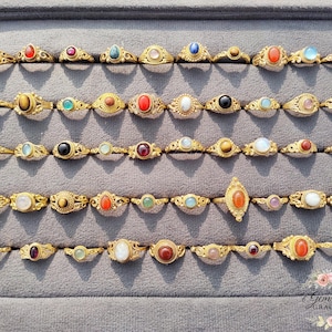 May include: A selection of gold-coloured rings, each set with a different gemstone, such as red, blue, and white. The rings are displayed on a grey surface, showcasing a variety of designs and colours. The gemstones are of various shapes.