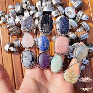 May include: A collection of gemstone rings with silver-colored bands. The rings feature various stone shapes and colors, including purple, black, blue, pink, and green. The rings are displayed on a hand, with many more rings in the background.