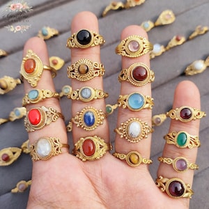 Puede incluir: Una colección de anillos dorados con diferentes engastes de piedras preciosas, incluyendo piedras negras, rojas, azules y blancas. Los anillos presentan diseños ornamentados de estilo filigrana y se muestran en dedos.