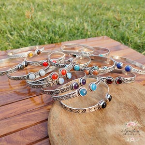 May include: A selection of silver bangles, each adorned with a different coloured gemstone, such as red, blue, and purple. The bangles are arranged on a wooden surface, with a blurred green backdrop.