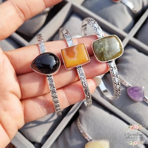 Op de afbeelding: Zilveren armbanden met verschillende edelstenen. De armbanden tonen een zwarte druppelvormige steen, een vierkante oranje steen en een vierkante groene steen. De zilveren banden hebben gedetailleerde patronen en worden gepresenteerd in een grijze sieradendoos.
