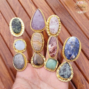 May include: A collection of gemstone rings with gold-colored settings. The rings feature various stone shapes and colors, including purple, green, blue, and brown. The rings are displayed on a hand against a wooden background.
