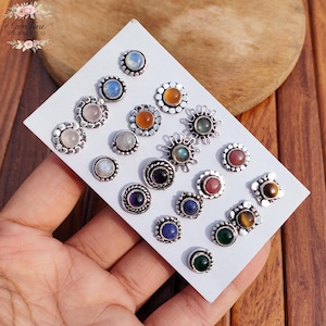 以下が含まれることがあります： 白いカードに展示された、宝石スタッドイヤリングのコレクション。イヤリングは、ムーンストーン、ローズクォーツ、アメジストなど、さまざまな宝石を銀色のセッティングで特徴としています。イヤリングは丸型と四角型で、さまざまな色があります。