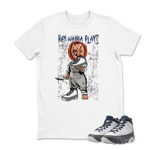 May include: White t-shirt with a graphic of a doll holding a sneaker and a knife, with the text "HEY WANNA PLAY?" in blue. A pair of white and gray high-top sneakers with blue accents are at the bottom.