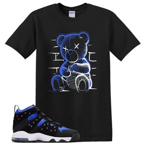 May include: A black short-sleeve t-shirt featuring a graphic of a seated teddy bear with X-shaped eyes in blue and white line art, paired with a black, blue, and white high-top sneaker with a visible air unit in the heel and a thick white sole.