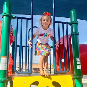 May include: A white short-sleeved t-shirt with the multicolored text "HADDIE" across the chest, paired with a vibrant, multi-colored crayon-patterned skirt, a bright orange hair bow, and tan sandals, standing on a colorful playground structure.