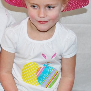 Puede incluir: Una persona joven con un sombrero de paja rosa brillante con ribete blanco y una camiseta blanca con un dise&ntilde;o de huevo de Pascua. La camiseta tiene un conejito y gr&aacute;ficos de huevos coloridos. La persona tambi&eacute;n lleva una falda colorida.