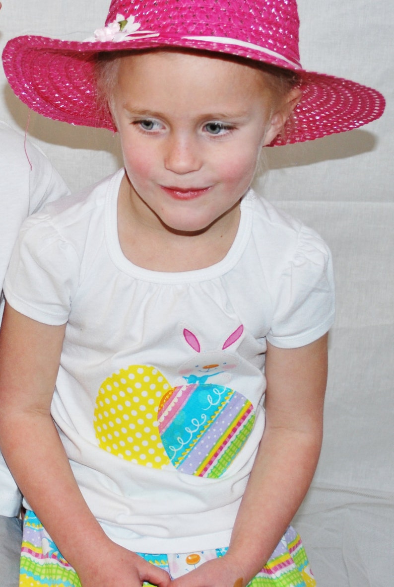 Puede incluir: Una persona joven con un sombrero de paja rosa brillante con ribete blanco y una camiseta blanca con un dise&ntilde;o de huevo de Pascua. La camiseta tiene un conejito y gr&aacute;ficos de huevos coloridos. La persona tambi&eacute;n lleva una falda colorida.