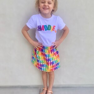 May include: A white short-sleeved t-shirt with the word CHADDIE printed in multicolored letters across the chest, paired with a gathered skirt featuring a repeating pattern of colorful pencils and tan leather sandals with cutouts.