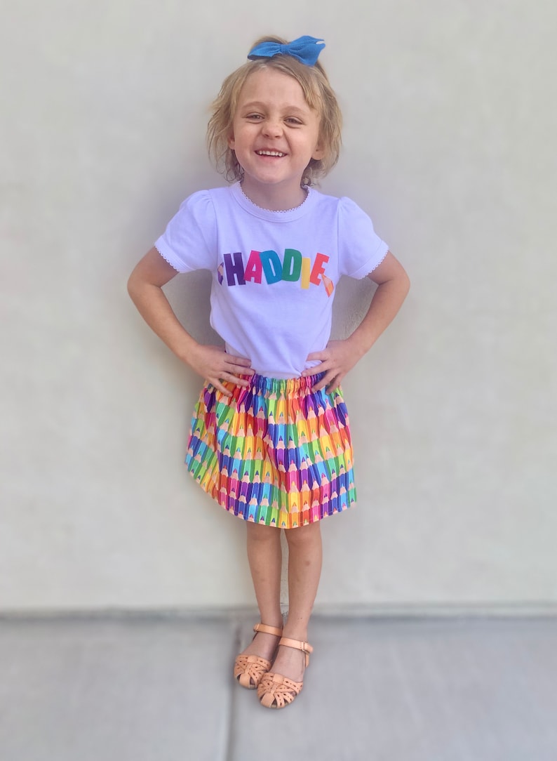 May include: A white short-sleeved t-shirt with the word CHADDIE printed in multicolored letters across the chest, paired with a gathered skirt featuring a repeating pattern of colorful pencils and tan leather sandals with cutouts.