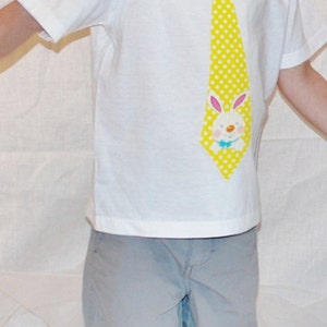 Puede incluir: Un ni&ntilde;o viste una camiseta blanca con una corbata amarilla de lunares y un dise&ntilde;o de conejito de dibujos animados. El ni&ntilde;o tambi&eacute;n lleva pantalones cortos grises.