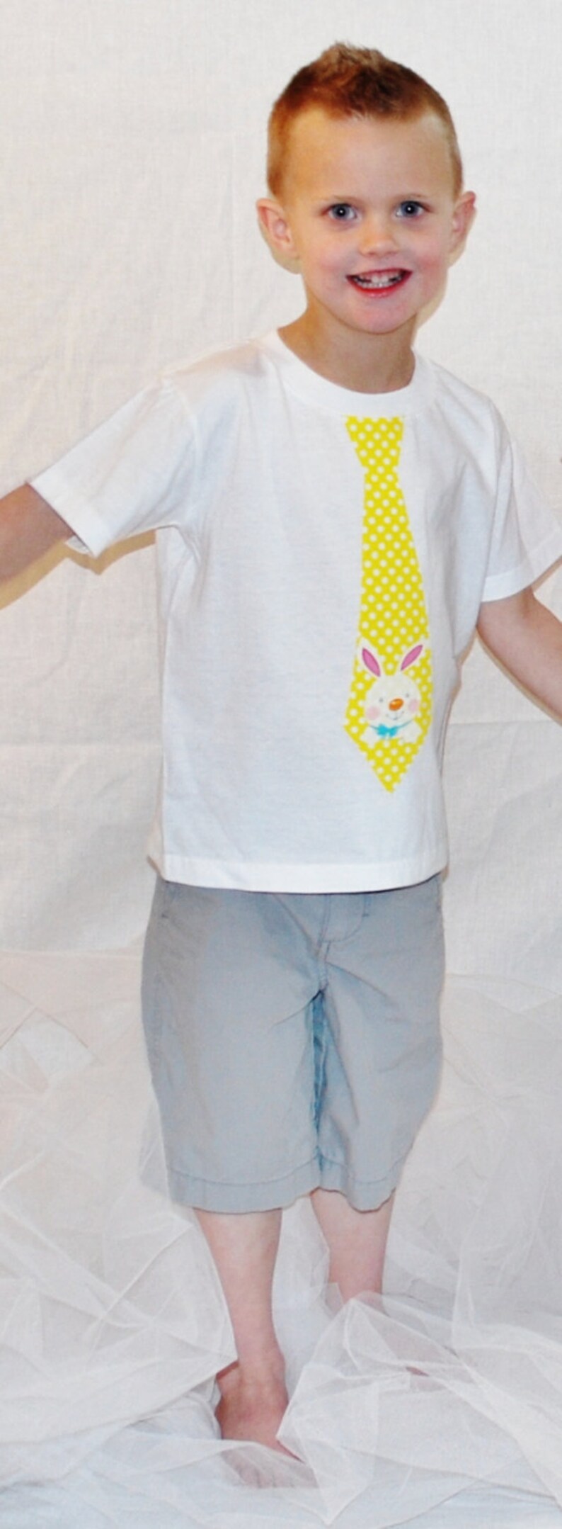 Puede incluir: Un ni&ntilde;o viste una camiseta blanca con una corbata amarilla de lunares y un dise&ntilde;o de conejito de dibujos animados. El ni&ntilde;o tambi&eacute;n lleva pantalones cortos grises.