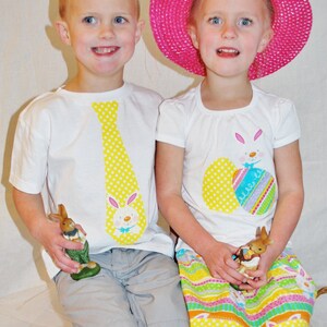 Puede incluir: Dos ni&ntilde;os con camisetas blancas con dise&ntilde;os de Pascua. Una camiseta tiene una corbata amarilla con un conejito, la otra un conejito y huevos decorados. La ni&ntilde;a lleva un sombrero rosa y una falda colorida, ambos sosteniendo figuras de conejitos.