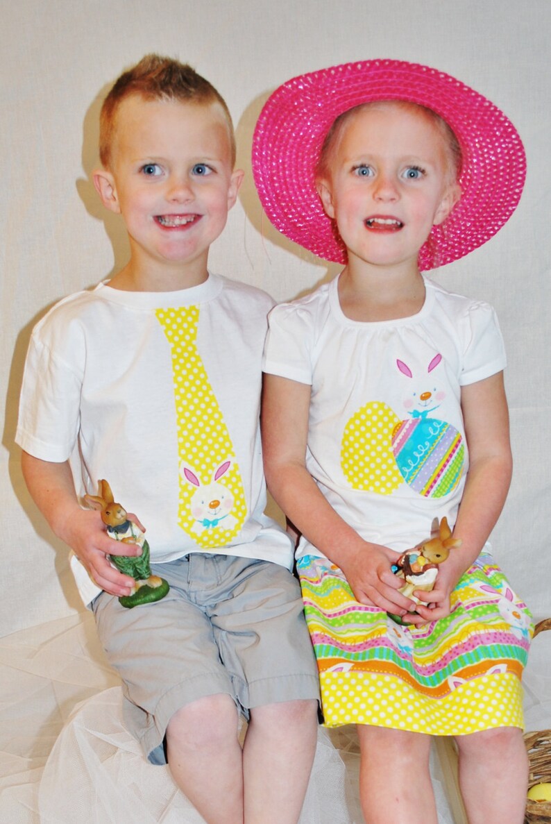 Puede incluir: Dos ni&ntilde;os con camisetas blancas con dise&ntilde;os de Pascua. Una camiseta tiene una corbata amarilla con un conejito, la otra un conejito y huevos decorados. La ni&ntilde;a lleva un sombrero rosa y una falda colorida, ambos sosteniendo figuras de conejitos.