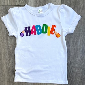 May include: A white short-sleeved t-shirt with a scalloped lace trim on the neckline and sleeves. The front features the name HADDIE in colorful block letters, flanked by a purple pencil on the left and an orange pencil on the right side.
