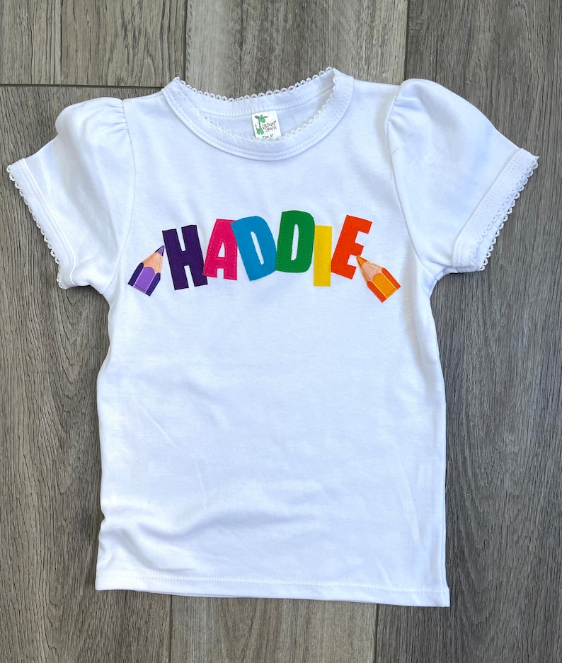 May include: A white short-sleeved t-shirt with a scalloped lace trim on the neckline and sleeves. The front features the name HADDIE in colorful block letters, flanked by a purple pencil on the left and an orange pencil on the right side.