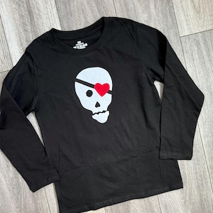 May include: Black long-sleeved t-shirt with a white skull and crossbones graphic. The skull has a red heart over one eye.