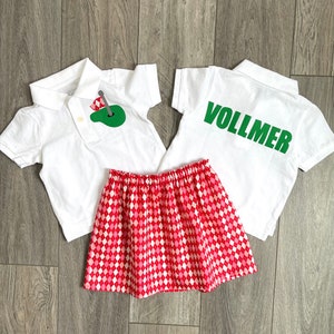 May include: Two white polo shirts with green text and a green golf ball graphic. One shirt says "VOLLMER". A red and white checkered skirt.