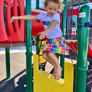 May include: A white short-sleeved t-shirt with the word "HADDIE" in multicolored block letters and a skirt featuring a repeating pattern of colorful crayons in shades of yellow, orange, red, purple, blue, and green, paired with tan sandals.