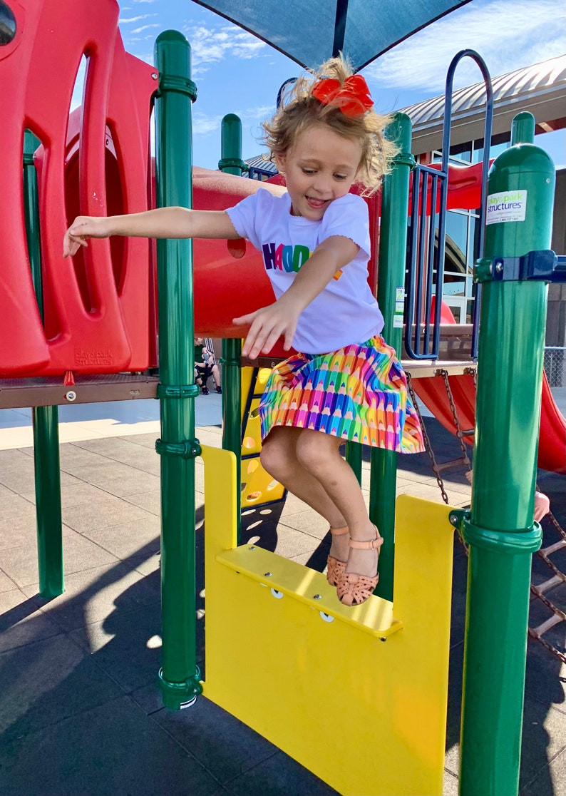 May include: A white short-sleeved t-shirt with the word "HADDIE" in multicolored block letters and a skirt featuring a repeating pattern of colorful crayons in shades of yellow, orange, red, purple, blue, and green, paired with tan sandals.