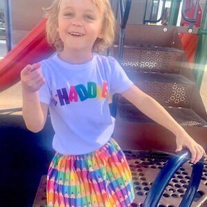 May include: A white short-sleeved t-shirt with the name HADDIE printed in multicolored block letters across the chest, paired with a gathered skirt featuring a repeating pattern of colorful sharpened pencils in various bright hues.