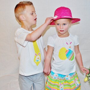 Puede incluir: Dos ni&ntilde;os con camisetas blancas con dise&ntilde;os de Pascua. Un ni&ntilde;o le pone un sombrero rosa a una ni&ntilde;a. La ni&ntilde;a lleva una falda colorida. Un ramo de flores con huevos de Pascua en el fondo sugiere una celebraci&oacute;n.
