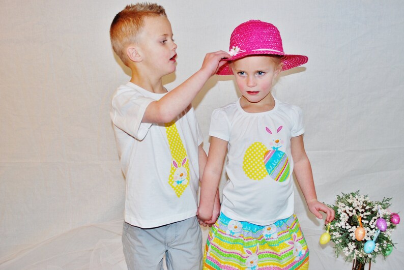 Puede incluir: Dos ni&ntilde;os con camisetas blancas con dise&ntilde;os de Pascua. Un ni&ntilde;o le pone un sombrero rosa a una ni&ntilde;a. La ni&ntilde;a lleva una falda colorida. Un ramo de flores con huevos de Pascua en el fondo sugiere una celebraci&oacute;n.