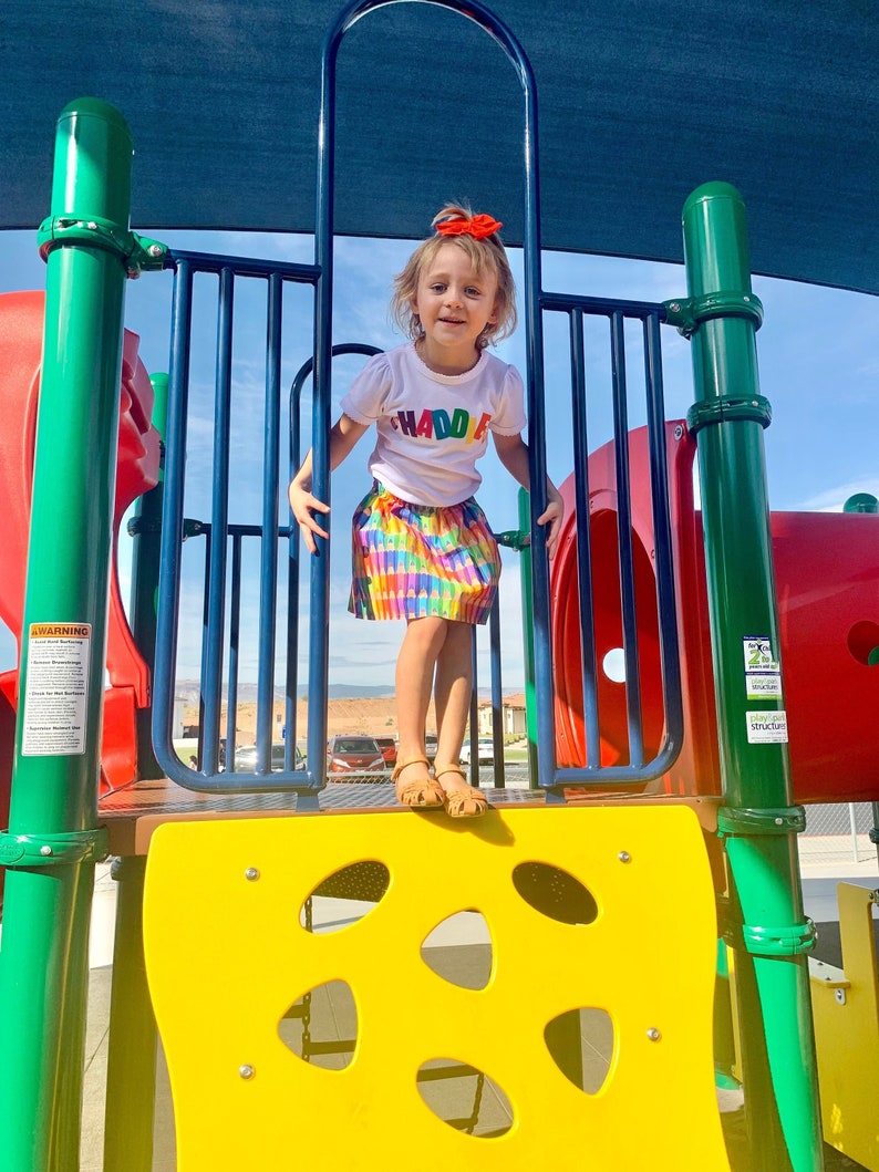 May include: A white short-sleeved t-shirt with the multicolored text "HADDIE" across the chest, paired with a vibrant, multi-colored crayon-patterned skirt, a bright orange hair bow, and tan sandals, standing on a colorful playground structure.