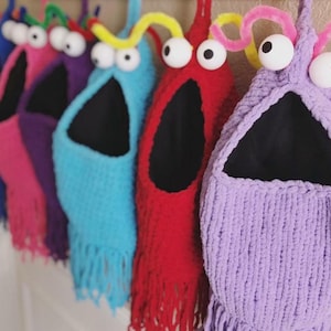 Può includere: Una fila di sacchi portaoggetti colorati fatti a mano a forma di mostro. Ogni sacco è di un colore diverso, tra cui blu, rosa, viola e rosso. Hanno grandi occhi mobili e frange sul fondo. I sacchi sono appesi su una superficie bianca.