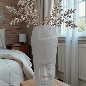 Op de afbeelding: Een witte keramische vaas met een gezichtsontwerp, met takken met lichtbruine bladeren. De vaas staat op een houten tafel, met een bed en een raam op de achtergrond. De vaas heeft een getextureerd oppervlak.