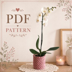 May include: A pink knitted plant pot with an orchid and white flowers. The pot is on a white lace doily. The words "PDF PATTERN" are in brown text above the plant.