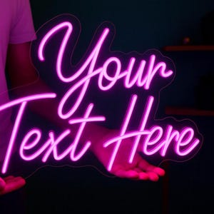May include: A neon sign in a cursive font that reads "Your Text Here" in bright pink. The sign is held up by a person, with the text glowing against a dark background. The sign is designed to be customized.