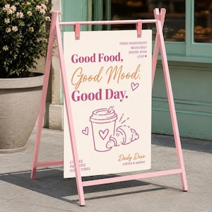Puede incluir: Un letrero de acera rosa en forma de A con un panel rectangular blanco que muestra el texto "Good Food, Good Mood, Good Day." y "Daily Dose COFFEE & BAKERY". El gráfico incluye un café para llevar y un cruasán en líneas finas.