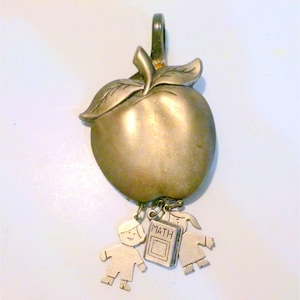 May include: A silver charm in the shape of an apple with a leaf and a stem. The apple has three charms hanging from it: two children and a book that says "MATH".