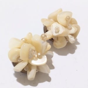 Puede incluir: Un par de pendientes de clip con un racimo de piedras de color blanquecino de forma irregular y pequeñas perlas redondas. Los pendientes tienen un diseño floral y están montados sobre clips de metal plateado.