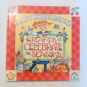 May include: A hardcover book titled "Crafts to Celebrate the Seasons" by Mary Engelbreit. The cover features a whimsical illustration of a girl in a sailor outfit holding a sign. The book has a red checkered border and decorative corner accents.