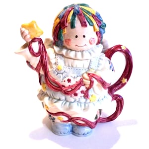 Puede incluir: Una tetera de cerámica con forma de muñeca de trapo con ropa de patchwork de colores, sosteniendo una estrella amarilla en una mano. La tetera tiene un asa de cinta roja y un patrón floral azul y blanco.