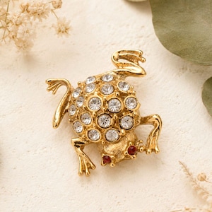 Puede incluir: Broche de rana dorada con detalles de cristal transparente y ojos rojos. El cuerpo de la rana está cubierto de cristales redondos y las patas están extendidas. Joya decorativa.