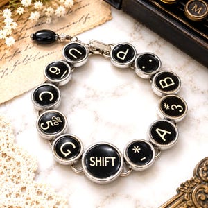 Könnte beinhalten: Ein kreisförmiges Armband aus einzelnen schwarzen Schreibmaschinentasten mit silberfarbenen Metallrändern. Die Tasten zeigen weiße aufgedruckte Zeichen, darunter Buchstaben, Zahlen, Symbole und das Wort SHIFT. Eine schwarze Perle hängt am Verschluss.