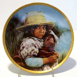 May include: A porcelain plate with a gold rim features a painting of a young girl wearing a straw hat and a colourful shawl. The girl is looking directly at the viewer with a serious expression. The background is a blurred landscape with a body of water.