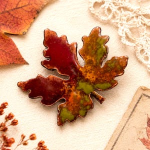 May include: A handcrafted leaf-shaped brooch with a copper border. The brooch features a blend of red, orange, and green enamel, resembling autumn foliage. The brooch is a decorative accessory, perfect for fall. The brooch is approximately 2 inches in length.