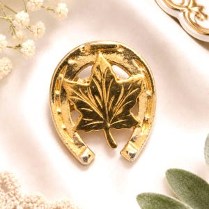 Broche vintage dourado com ferradura da sorte e folha de bordo.