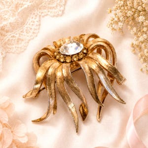 May include: A vintage gold-tone brooch with a large, clear rhinestone center surrounded by smaller rhinestones. The brooch has a stylized floral design with long, curved petals. The background is a soft, cream-colored fabric.
