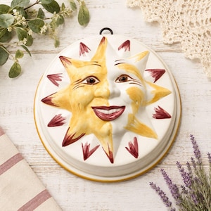 May include: A round ceramic wall plaque featuring a sculpted sun face with a split design. One half is painted in yellow with brown accents, while the other half is white. The sun has a smiling face, pointed rays, and a dark metal hanging loop.